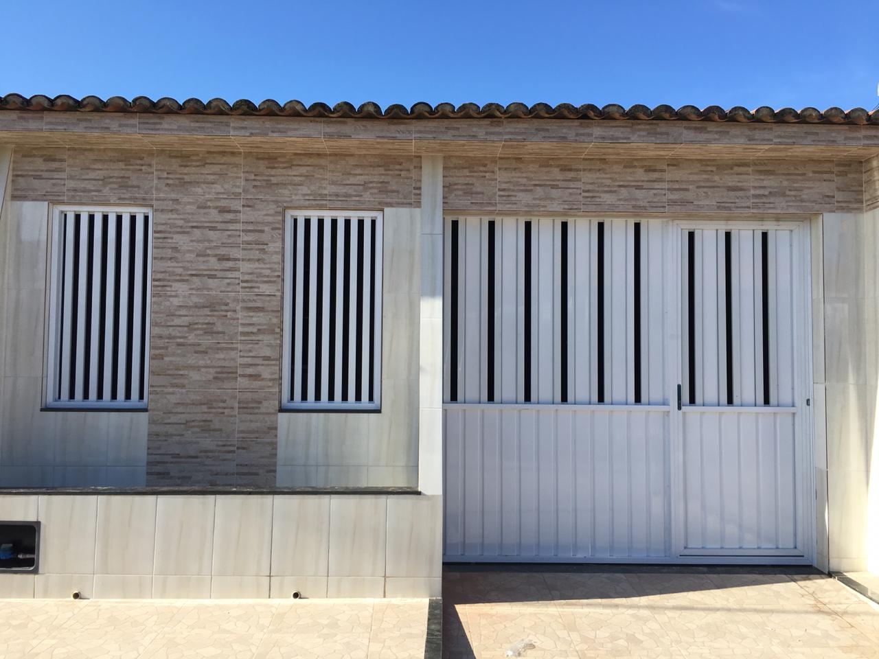 Portão em alumínio branco, de correr com porta social acoplada. Modelo: fechado embaixo com lambril liso quadrado, em cima vasado com lambril e tubos, com grades fixas vasadas de tubos.